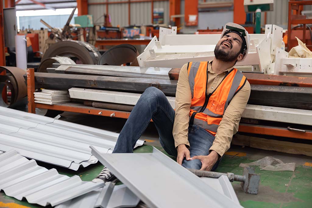 Injured industrial worker holds leg while leaning against construction pieces